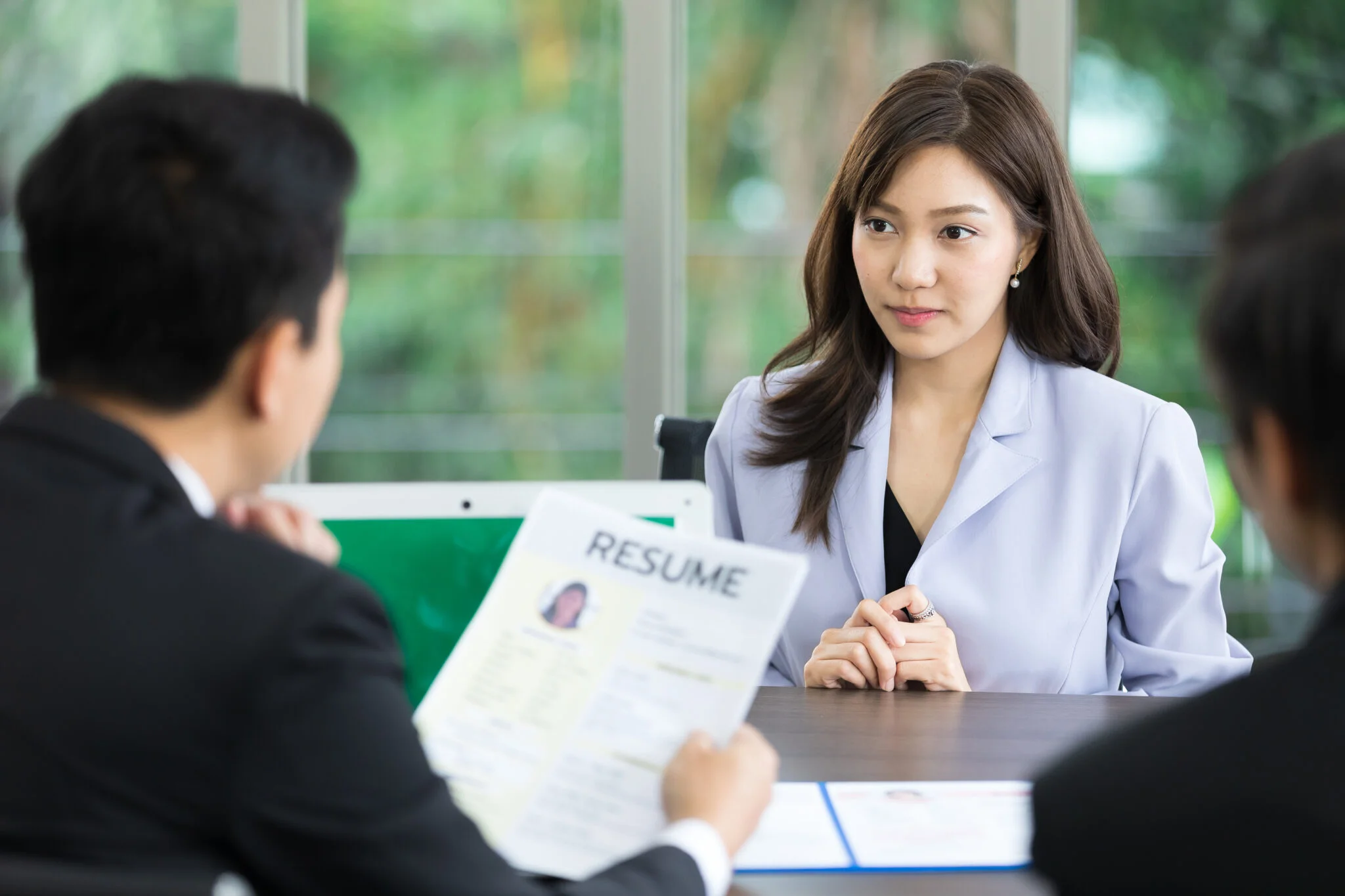 A job candidate in a light blue blazer is seated across from two interviewers at a table, with one interviewer holding and reviewing a resume.